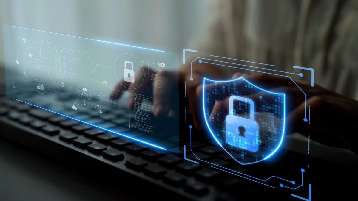A user's hands are visible typing on a computer keyboard, with a glowing blue digital security shield displaying a lock icon prominently on the right, signifying online safety, data encryption, and protection against cyber threats.